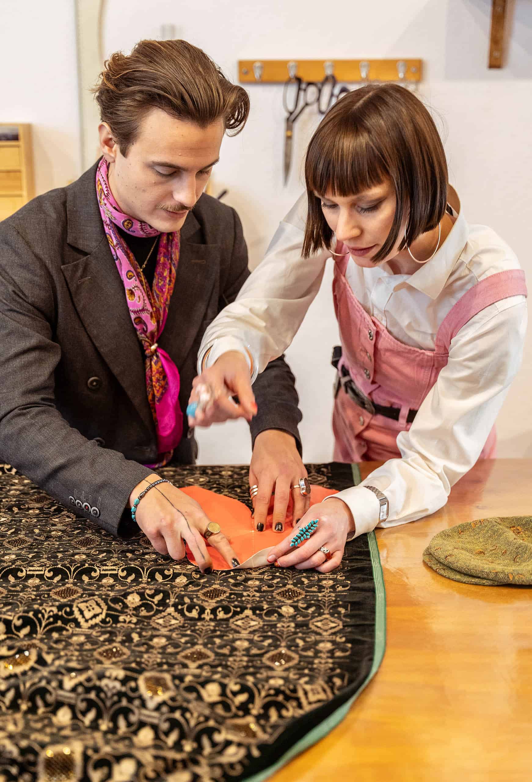 Chic:Leger | Zwei Modedesigner*innen beim präzisen Zuschneiden von Stoff an einem Werktisch