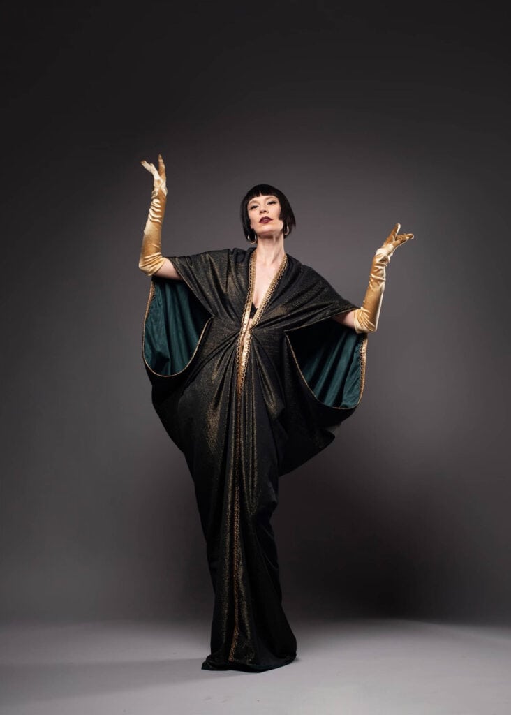Frau in elegantem, dunklem Cape-Kleid mit goldenen Handschuhen und glamouröser Pose