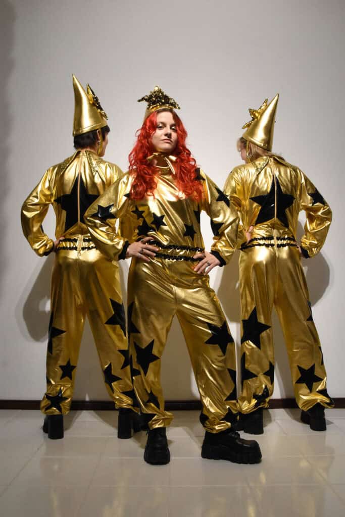 Drei Performer*innen in goldenen Overalls mit schwarzen Sternen und spitzen Hüten