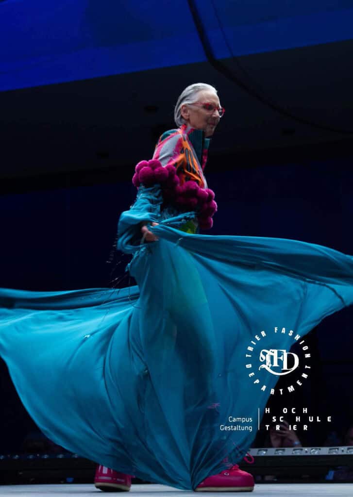 Älteres Model in ausladendem, türkisfarbenem Kleid mit pinken Applikationen